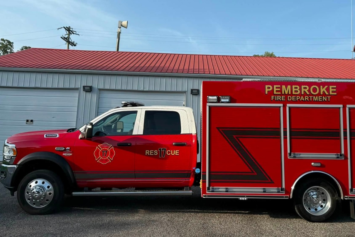 Red Pembroke Fire Department rescue truck parked outside a garage, with a blue sky above and a red roof building in the background.
