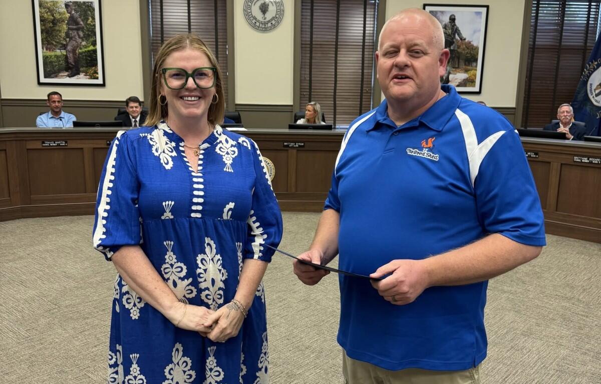 Smiling woman in a blue patterned dress and green glasses stands next to a man in a blue polo who holds a certificate in a council chamber.