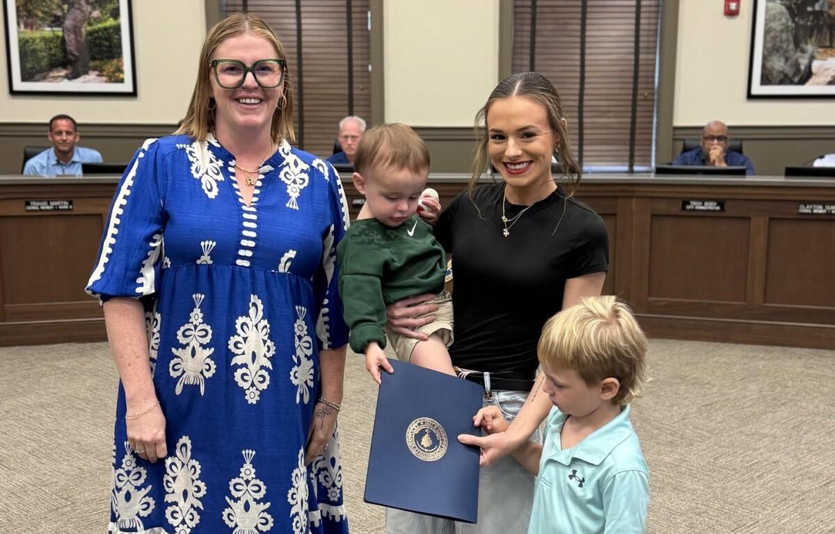Two women stand in a council chamber with a small child and a young boy holding a blue certificate folder, smiling at the camera.