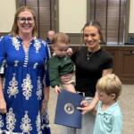Two women stand in a council chamber with a small child and a young boy holding a blue certificate folder, smiling at the camera.