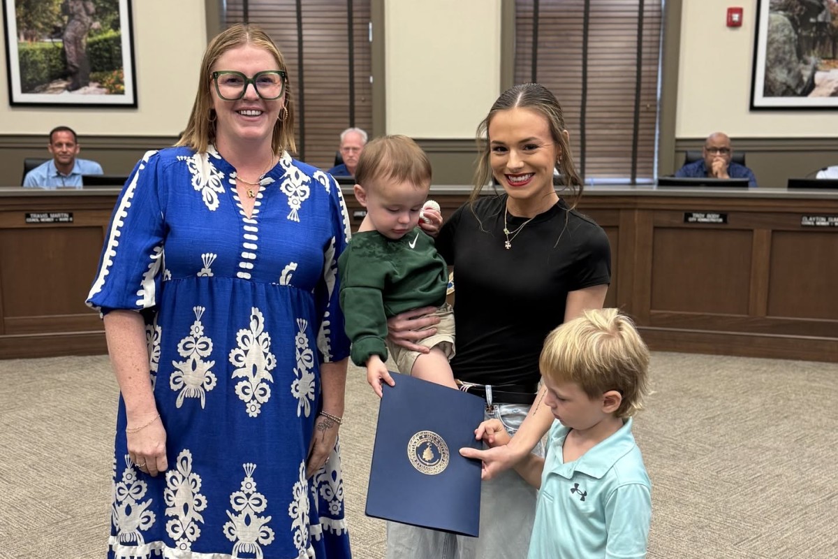 Two women stand in a council chamber with a small child and a young boy holding a blue certificate folder, smiling at the camera.