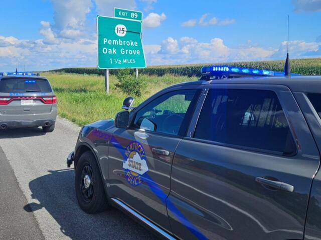 Two Kentucky State Police SUVs with blue lights parked on a rural highway next to an exit sign for Pembroke and Oak Grove, 1½ miles away.