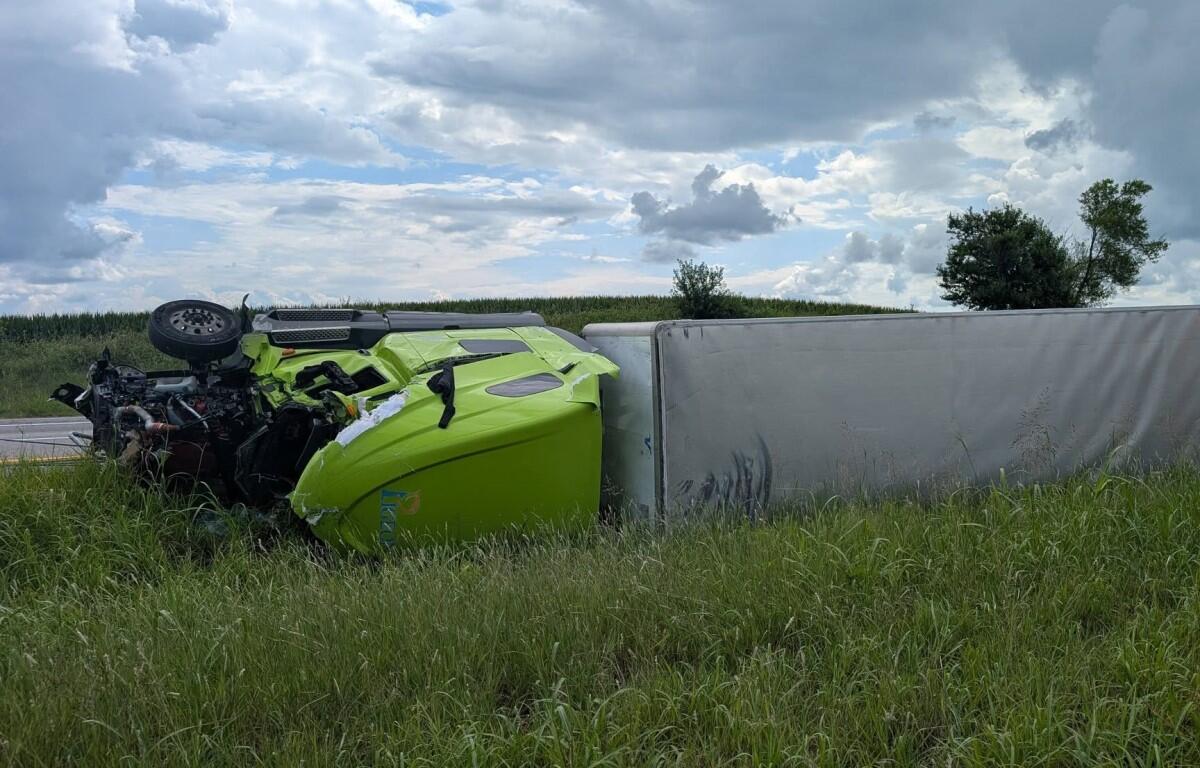 Lime-green sports car overturned on its roof against a gray trailer on a grassy roadside, engine exposed after a crash.