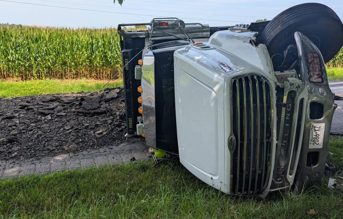 White semi-truck tipped onto its side on a rural road beside a cornfield, front grille facing the camera.