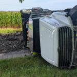 White semi-truck tipped onto its side on a rural road beside a cornfield, front grille facing the camera.
