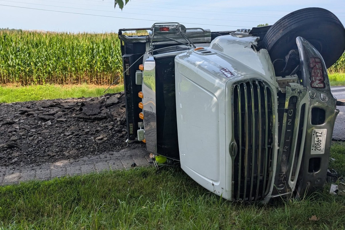 White semi-truck tipped onto its side on a rural road beside a cornfield, front grille facing the camera.