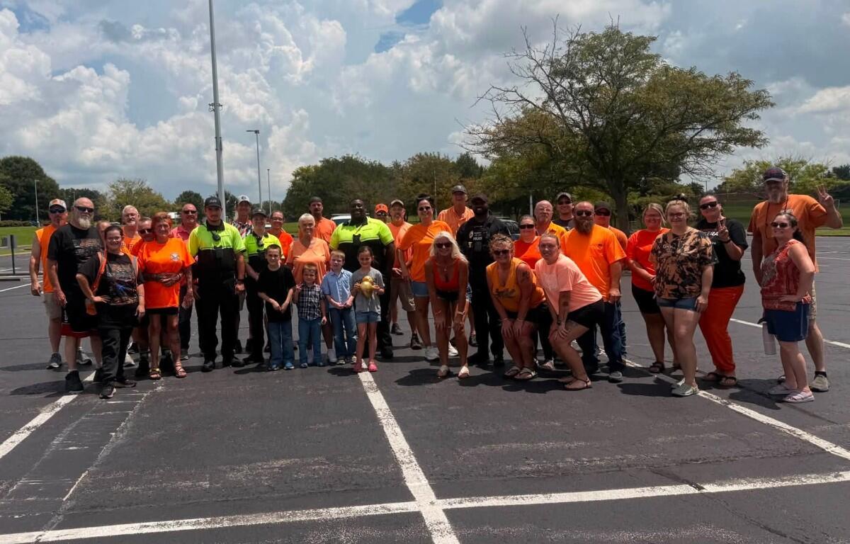 Large group of people in orange shirts posing together in a parking lot on a sunny day.