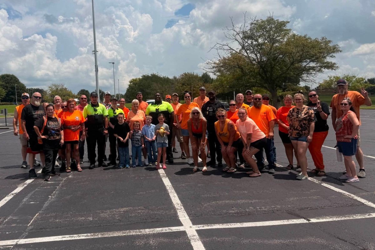 Large group of people in orange shirts posing together in a parking lot on a sunny day.