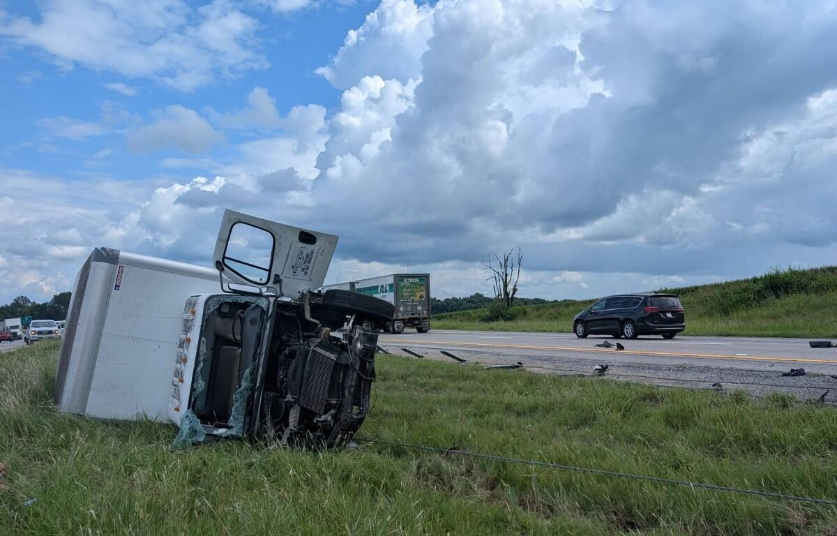 Overturned semi-truck on grassy shoulder with crushed cab and debris on the road, cloudy sky above.
