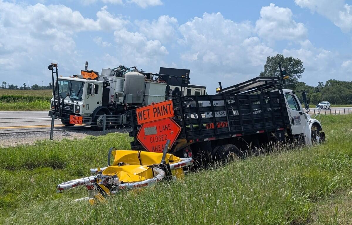Two large trucks on a rural roadside with orange 'WET PAINT' signs and a 'LANE CLOSED AHEAD' sign; a yellow barrier lies in tall grass nearby.
