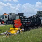 Two large trucks on a rural roadside with orange 'WET PAINT' signs and a 'LANE CLOSED AHEAD' sign; a yellow barrier lies in tall grass nearby.