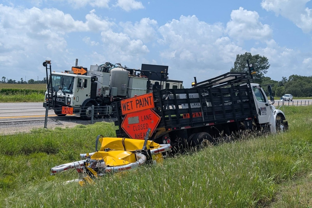 Two large trucks on a rural roadside with orange 'WET PAINT' signs and a 'LANE CLOSED AHEAD' sign; a yellow barrier lies in tall grass nearby.