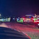 Nighttime scene with sheriff and police vehicles parked in a residential yard, officers and bystanders near a central car, red/blue lights flashing.