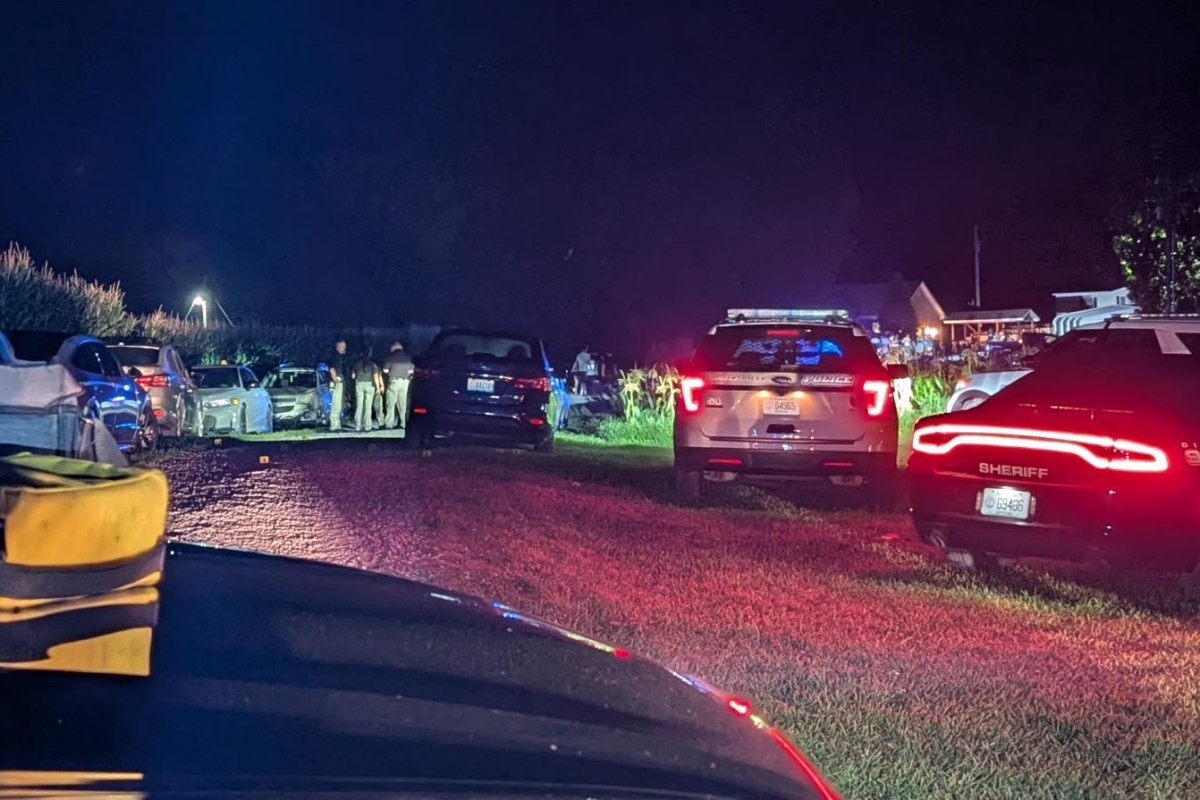Nighttime scene with sheriff and police vehicles parked in a residential yard, officers and bystanders near a central car, red/blue lights flashing.