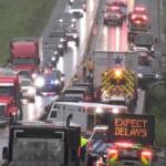 Highway scene with emergency vehicles and a bright sign that reads 'EXPECT DELAYS' amid a traffic backup on a wet road