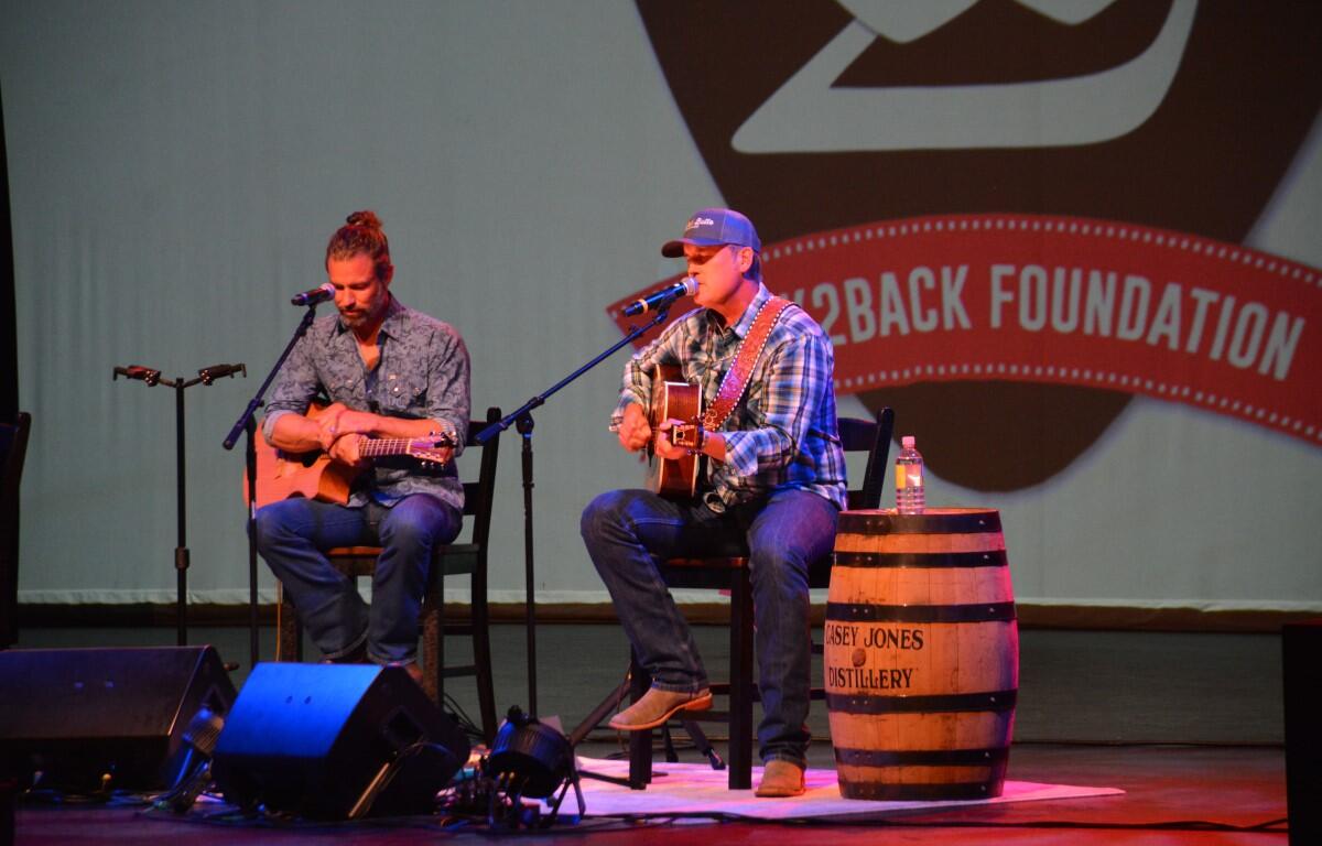 Two male musicians perform a live acoustic set on stage, each with an acoustic guitar and a microphone.