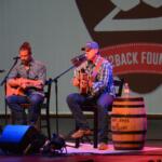 Two male musicians perform a live acoustic set on stage, each with an acoustic guitar and a microphone.