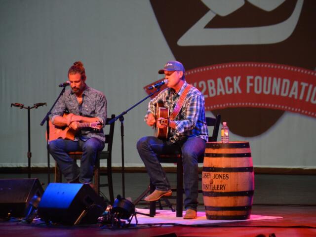Two male musicians perform a live acoustic set on stage, each with an acoustic guitar and a microphone.