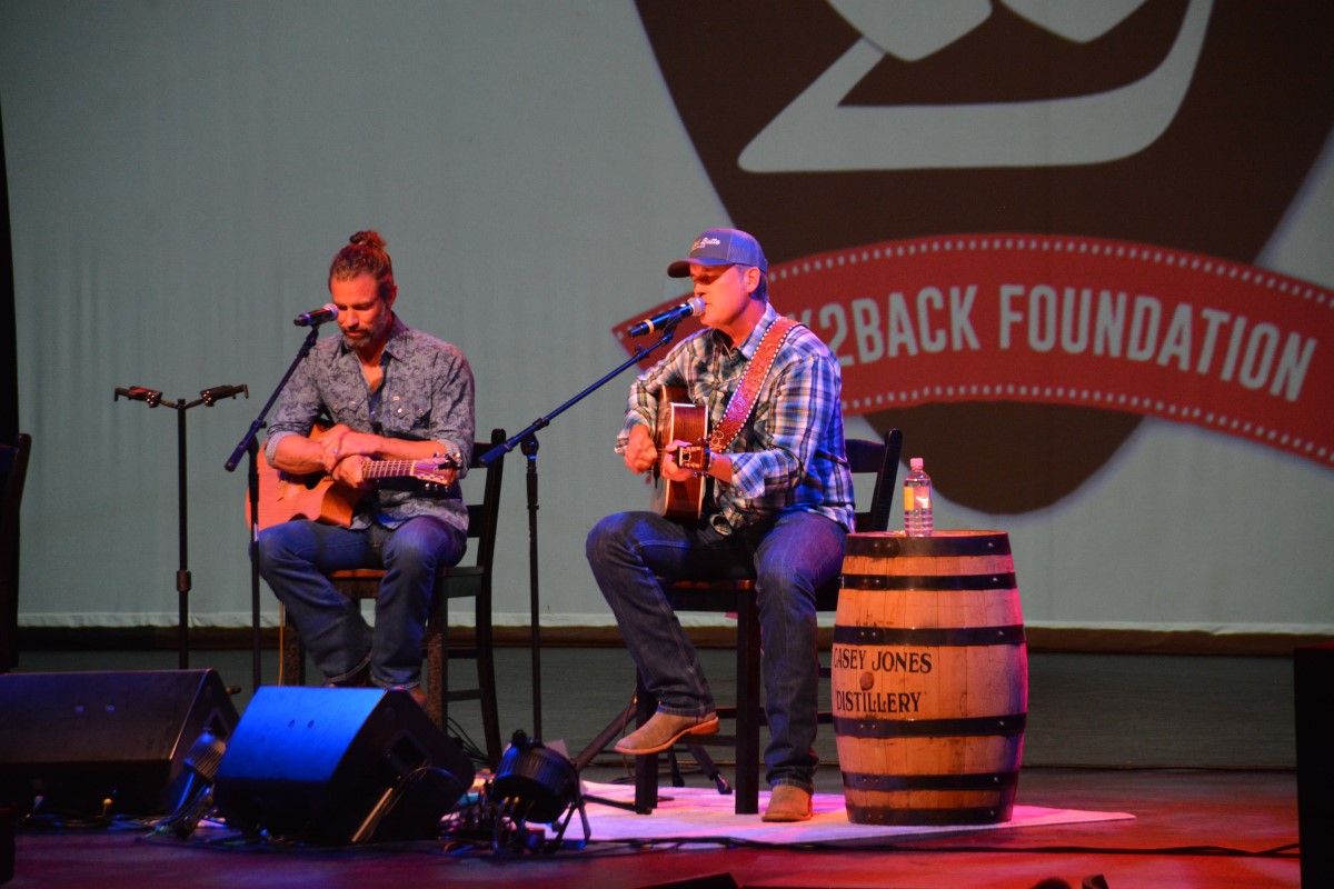 Two male musicians perform a live acoustic set on stage, each with an acoustic guitar and a microphone.