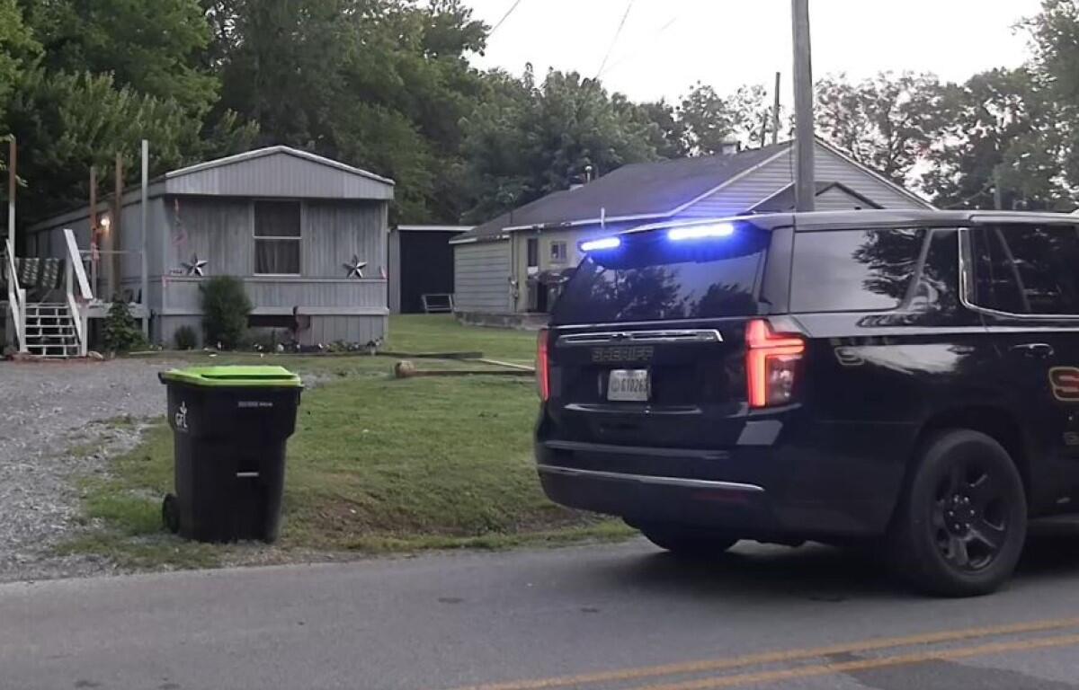 Sheriff SUV with blue lights on a residential street near mobile homes and a green trash bin in the yard.