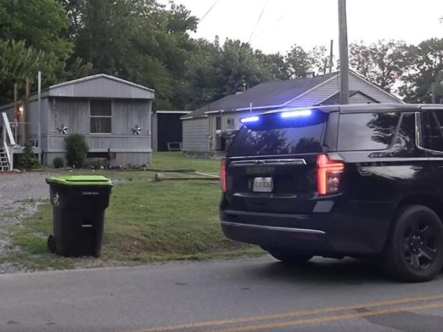 Sheriff SUV with blue lights on a residential street near mobile homes and a green trash bin in the yard.