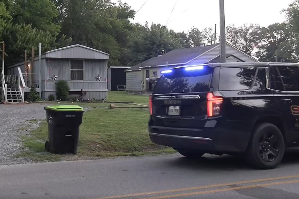 Sheriff SUV with blue lights on a residential street near mobile homes and a green trash bin in the yard.