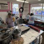 Concession booth with volunteers cooking and assembling food on foil-covered counter inside a pink-walled tent.