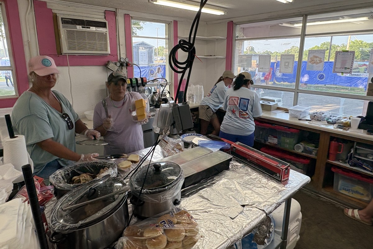 Concession booth with volunteers cooking and assembling food on foil-covered counter inside a pink-walled tent.