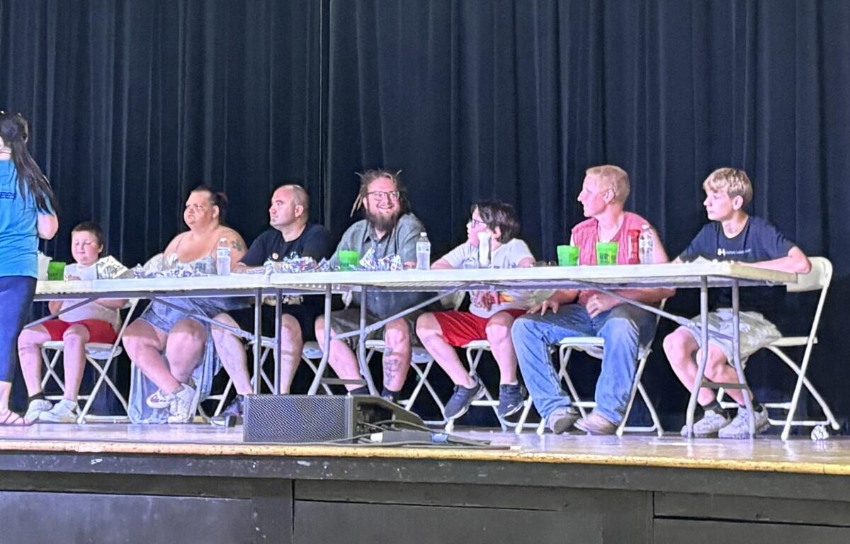 Several people sit in a row at a long folding table on a stage, eating snacks and drinking, with a dark curtain behind them.