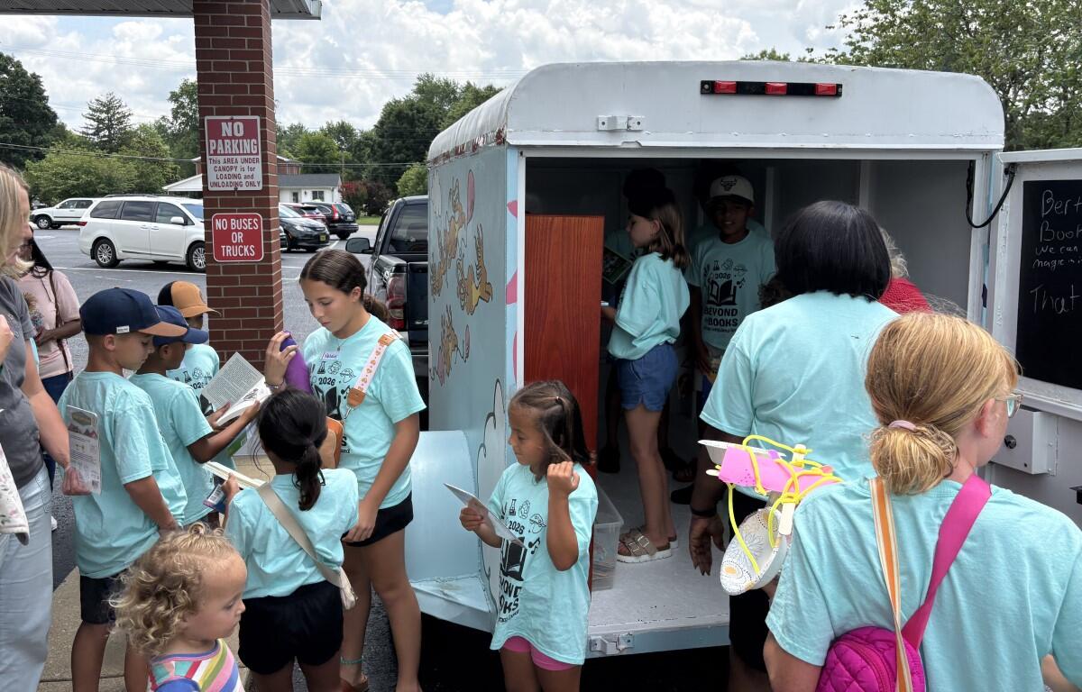 Children in turquoise shirts gather around a bookmobile, selecting and reading books from the mobile library truck in a parking lot.