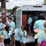 Children in turquoise shirts gather around a bookmobile, selecting and reading books from the mobile library truck in a parking lot.