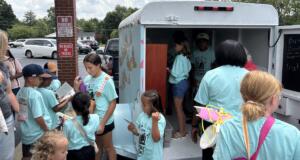 Children in turquoise shirts gather around a bookmobile, selecting and reading books from the mobile library truck in a parking lot.