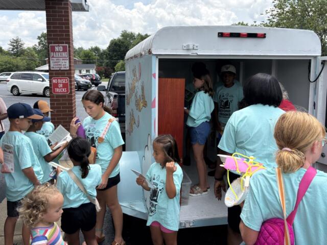Children in turquoise shirts gather around a bookmobile, selecting and reading books from the mobile library truck in a parking lot.