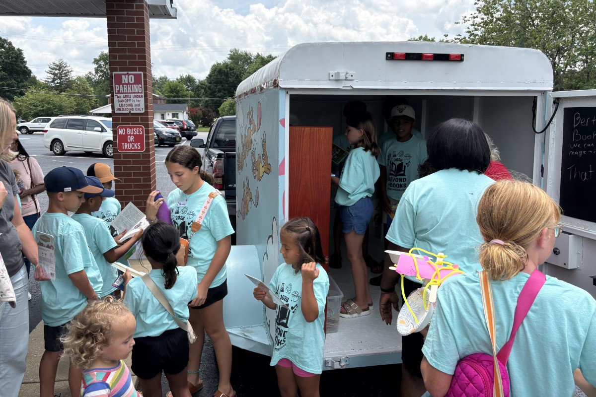 Children in turquoise shirts gather around a bookmobile, selecting and reading books from the mobile library truck in a parking lot.