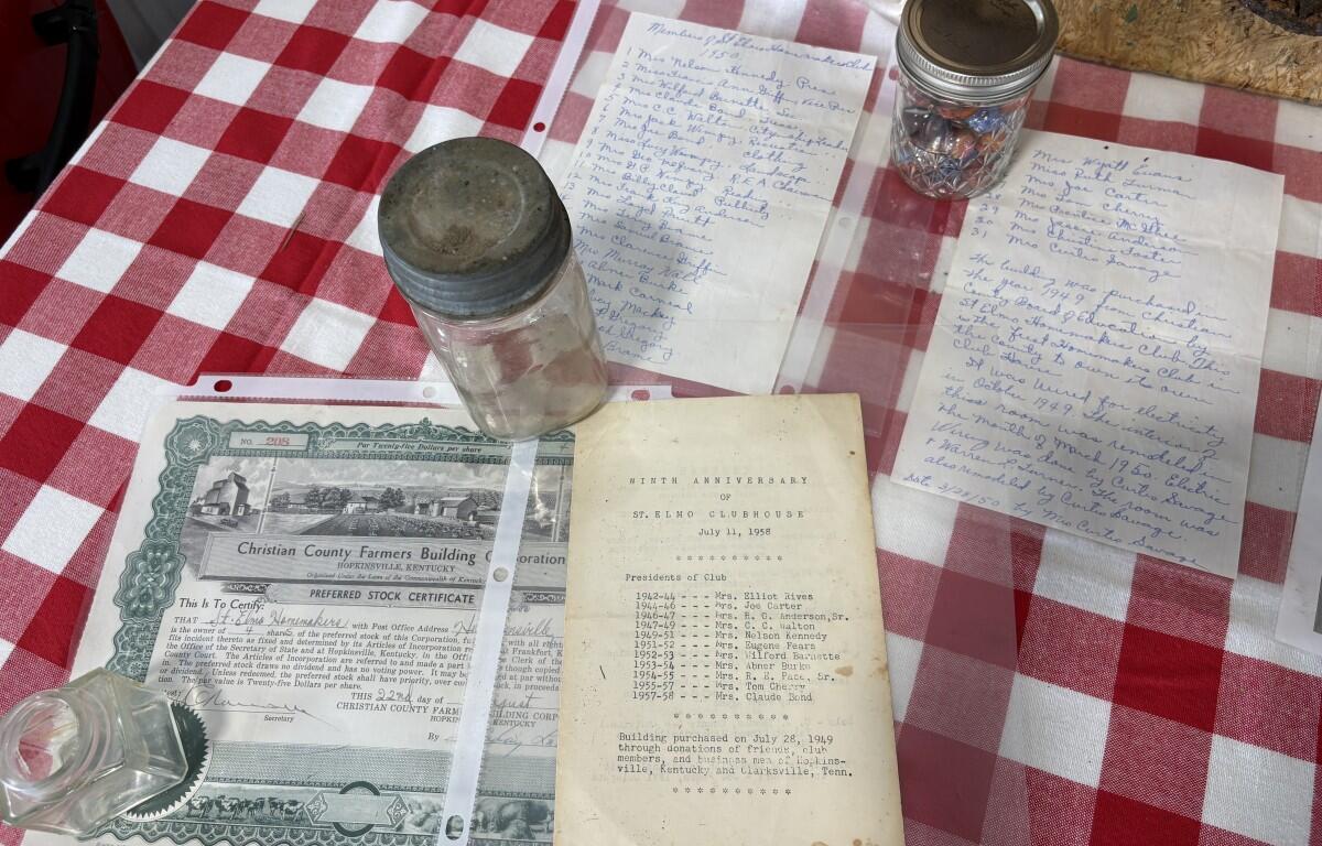 Display of vintage documents on a red-and-white checkered table: a booklet titled Ninth Anniversary of St. Elmo Clubhouse (1958), a stock certificate for Christian County Farmers Building & Loan, handwritten notes, a glass jar, and a metal canister.