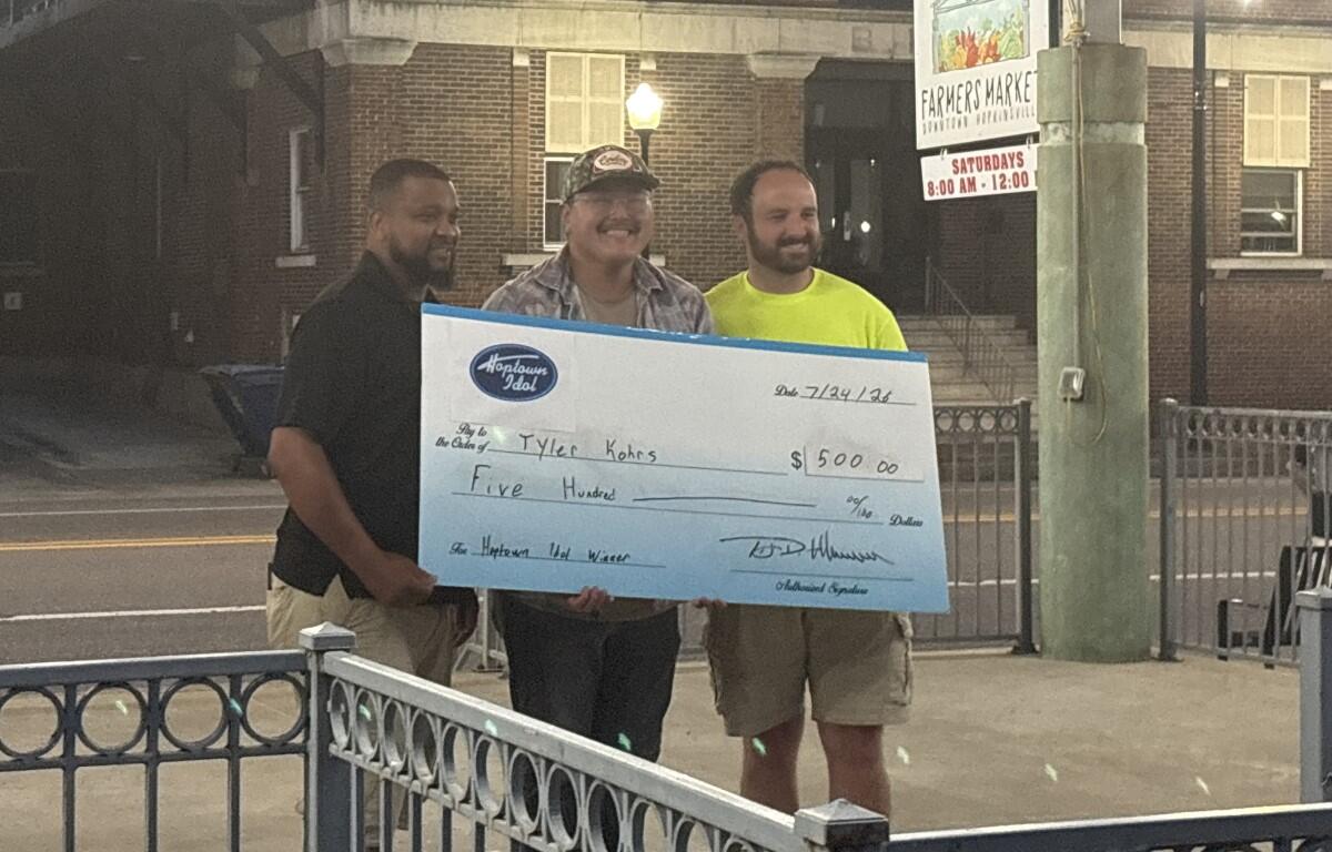 Three men stand on a sidewalk at night, holding a large ceremonial check for $5,000 in front of a brick building and a farmers market sign behind them.