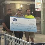 Three men stand on a sidewalk at night, holding a large ceremonial check for $5,000 in front of a brick building and a farmers market sign behind them.