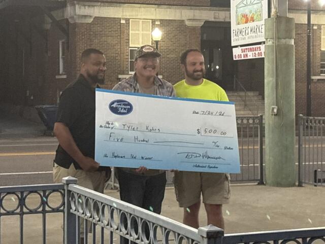 Three men stand on a sidewalk at night, holding a large ceremonial check for $5,000 in front of a brick building and a farmers market sign behind them.