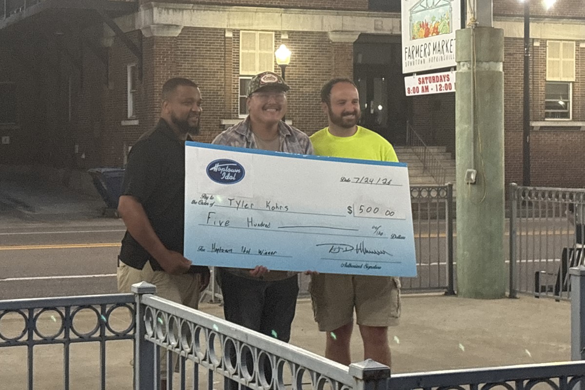Three men stand on a sidewalk at night, holding a large ceremonial check for $5,000 in front of a brick building and a farmers market sign behind them.