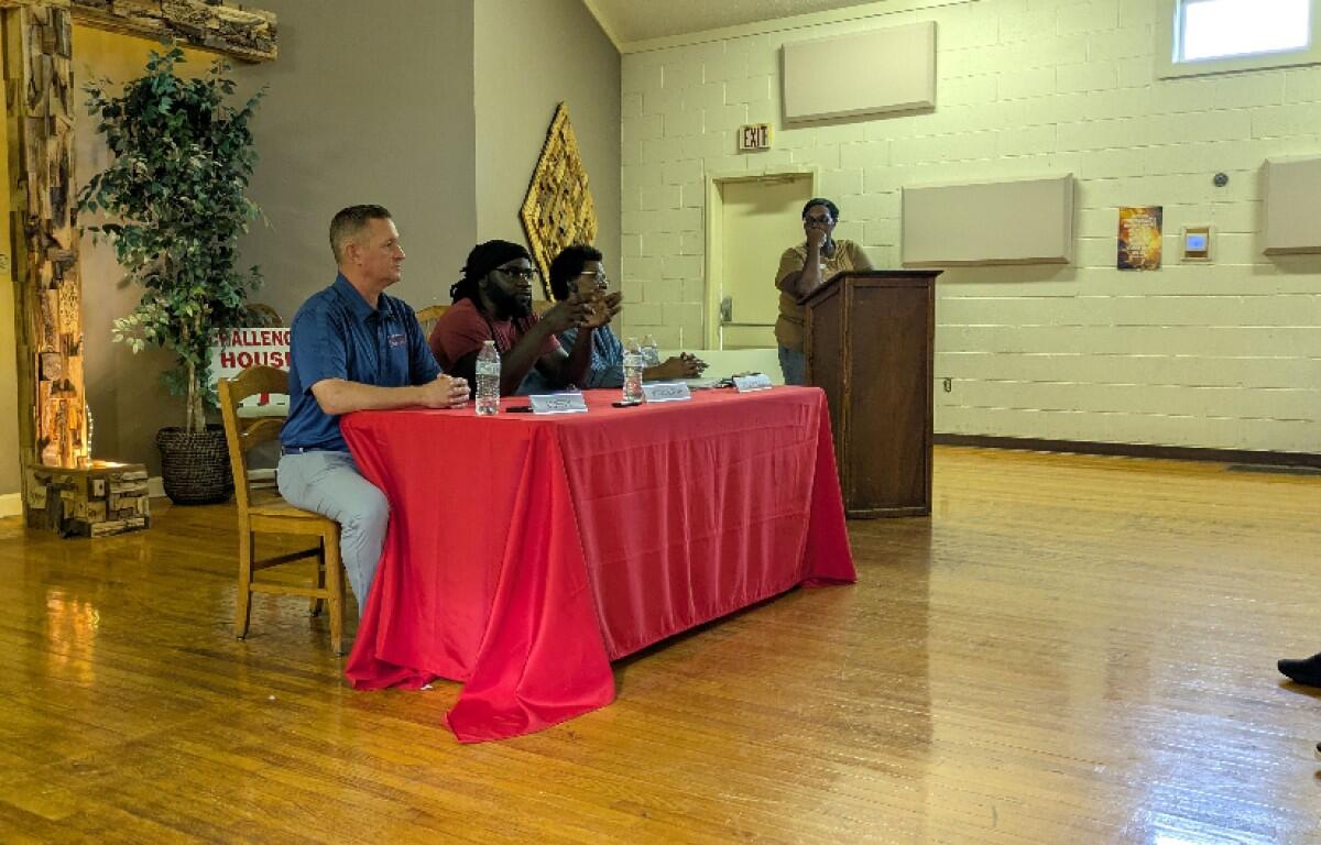 Panel of four people seated at a red tablecloth-covered table, one person at a podium speaking in a community room.