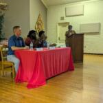 Panel of four people seated at a red tablecloth-covered table, one person at a podium speaking in a community room.