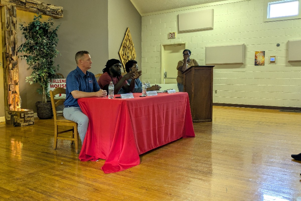 Panel of four people seated at a red tablecloth-covered table, one person at a podium speaking in a community room.