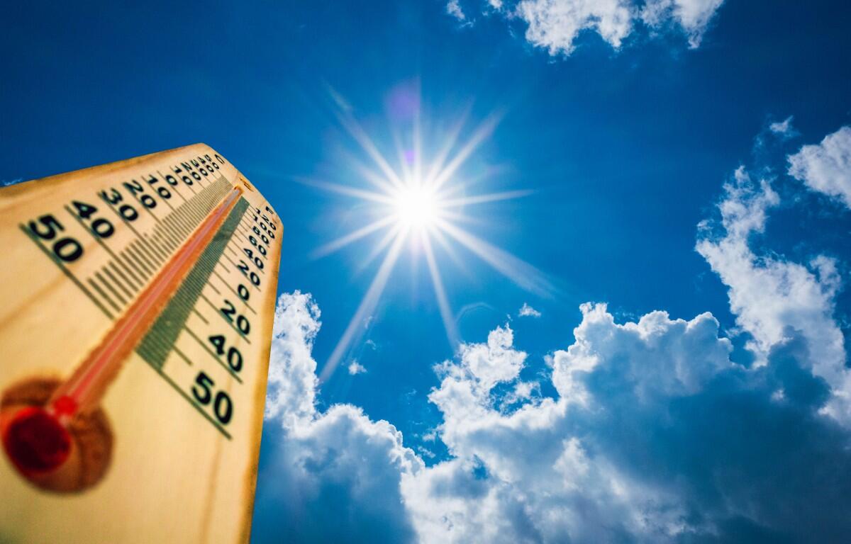 Wooden thermometer angled up toward a bright sun in a blue sky with clouds, conveying hot weather.