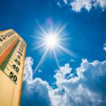 Wooden thermometer angled up toward a bright sun in a blue sky with clouds, conveying hot weather.