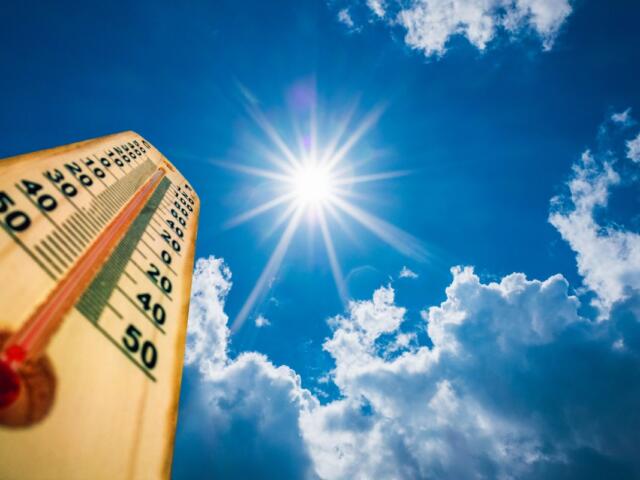 Wooden thermometer angled up toward a bright sun in a blue sky with clouds, conveying hot weather.