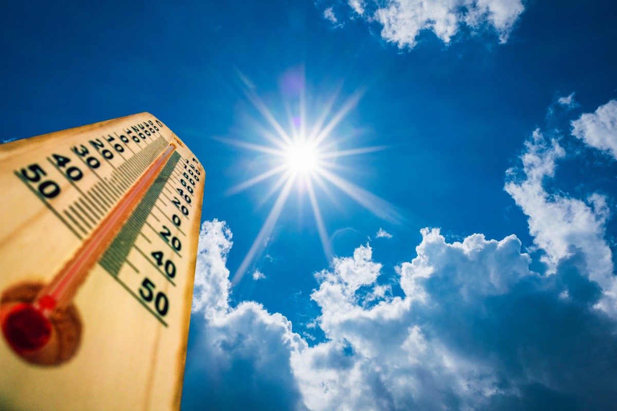 Wooden thermometer angled up toward a bright sun in a blue sky with clouds, conveying hot weather.