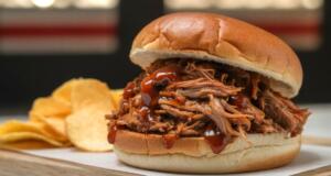 Pulled pork barbecue sandwich on a soft bun with dark sauce, served on a wooden board with chips on the side.