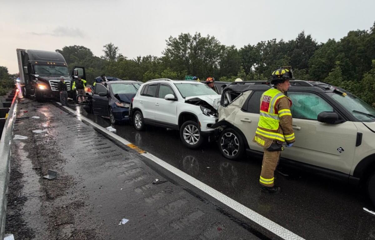 Multi‑vehicle crash on a rainy highway: a white SUV and a beige SUV show front‑end damage, backed by a dark blue car, with a tow truck arriving and firefighters in reflective gear on the scene.