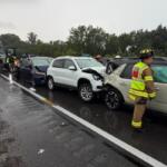 Multi‑vehicle crash on a rainy highway: a white SUV and a beige SUV show front‑end damage, backed by a dark blue car, with a tow truck arriving and firefighters in reflective gear on the scene.