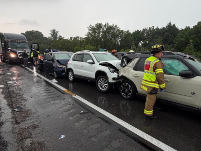 Multi‑vehicle crash on a rainy highway: a white SUV and a beige SUV show front‑end damage, backed by a dark blue car, with a tow truck arriving and firefighters in reflective gear on the scene.